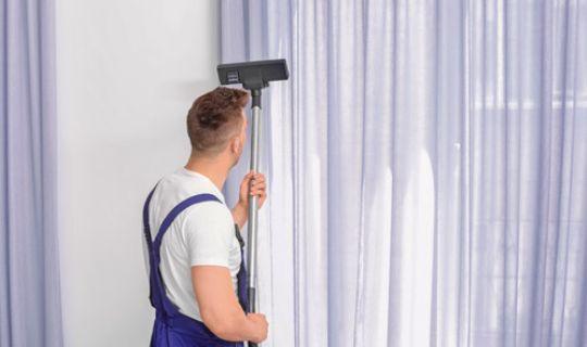 a professional using the wand of a hoover to clean hanging curtains