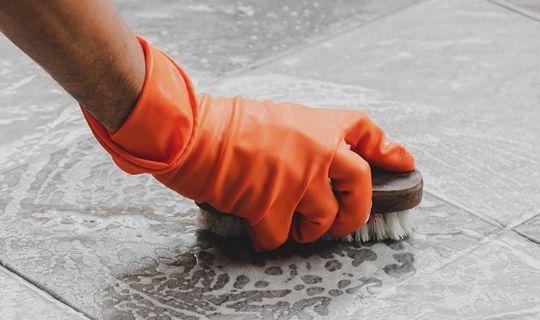 a cleaner's hand wearing a protective glove, scrubbing tiles with a brush and working a cleaning solution onto them