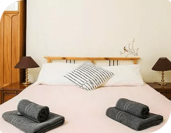 Picture of a bed with pink bedding, one striped pillow and two folded grey towels on it, side lamps and a wardrobe.