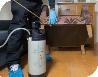 A pest controller, wearing protective gear sprays insecticide on the floor near a couch.