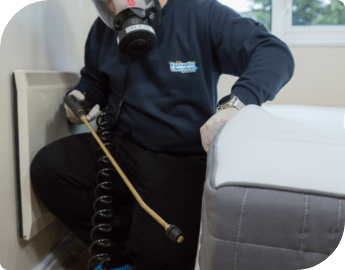 Pest expert in protective gear stands next to a bed and prepares to spray for insects.