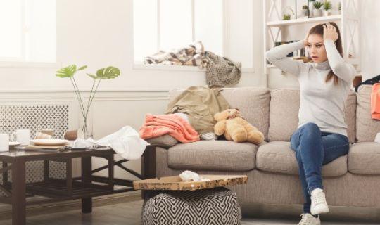 an overwhelmed woman sitting on her sofa and a messy room with clothes and clutter all around