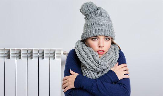 a woman wearing a scarf and a winter hat being cold and standing next to a radiator