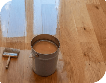 A bucket of lacquer and a paintbrush placed on a wooden floor, ready for floor refinishing service.