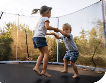 Two children holding hands and jumping on a trampoline outdoors.