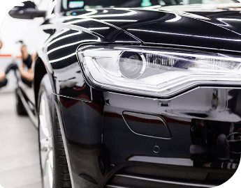 Close-up of a black car's front headlight and a professional in the background who is performing a refinement job