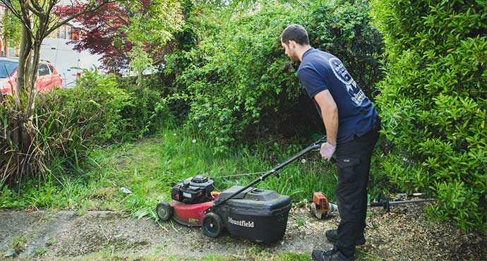 The image shows a gardener who is trimming an overgrown shrub. He is using a power tool with an extendable pole to remove the overgrown branches. The trimmings are collected in green waste bags.
