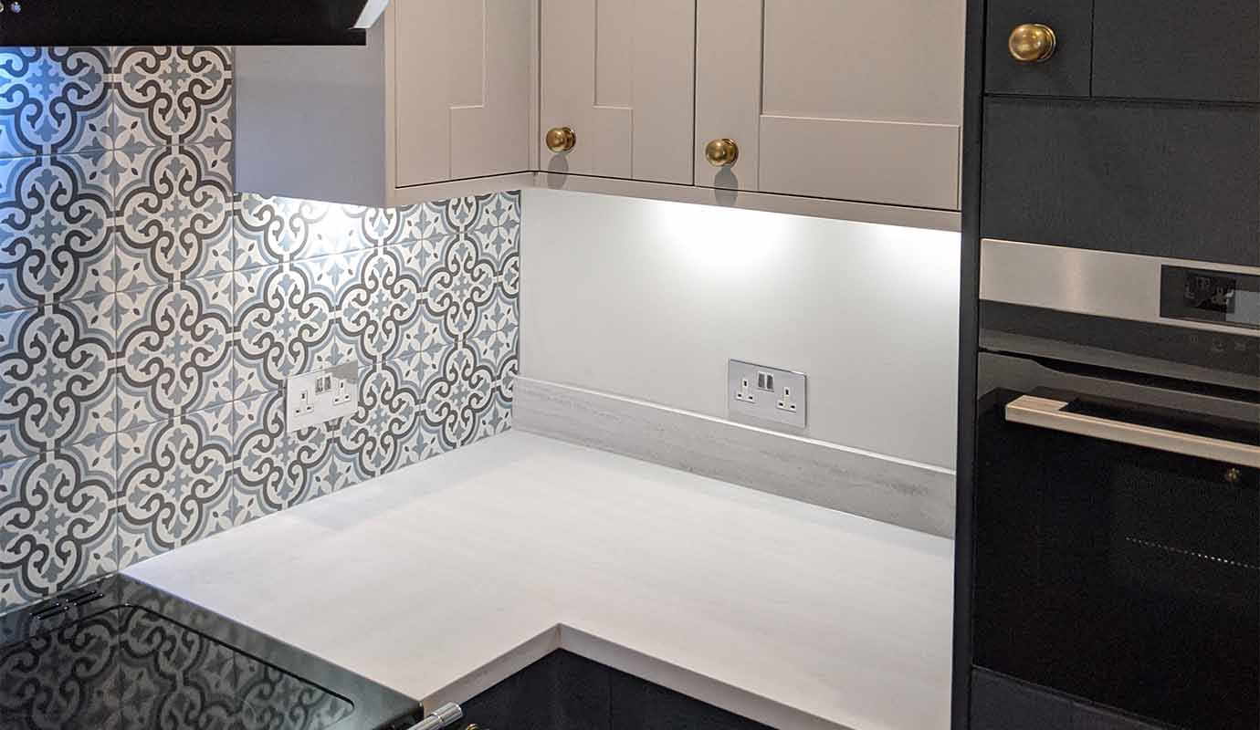 Modern kitchen corner with patterned backsplash, beige cabinets, black oven, and extractor hood.
