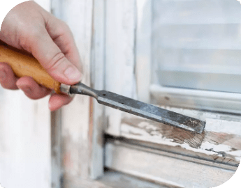 Close-up of the hand of a window repair technician who is scraping old paint from a wooden window frame with a chisel.