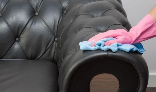 a cleaner's hand wearing a yellow protective glove using a blue microfibre cloth to wipe down a leather sofa