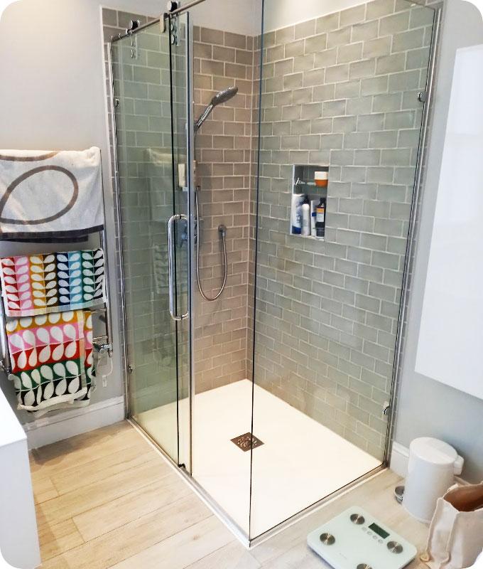 A bathroom corner with a shower cabin. This area of the bathroom appears perfectly clean and neat. There are fresh colourful towels placed on the towel racks. There is a digital weight scale on the wooden floor of the bathroom next to a rubbish bin. The shower cabin is stocked with shampoo, shower gel, and other bathing products.