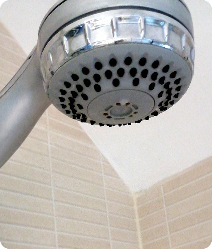 A close-up shot of the same chrome shower head in a bathroom fixed on a tiled wall. The shower head has been cleaned and descaled. The build-up of minerals has been removed and the chrome shower head looks polished and shiny.