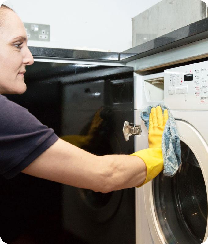A Fantastic Services domestic cleaner who is kneeling down in front of a white washing machine that is positioned in a compartment under a ceramic kitchen countertop. She is wearing yellow latex gloves and wiping the front of the washing machine with a cleaning cloth.