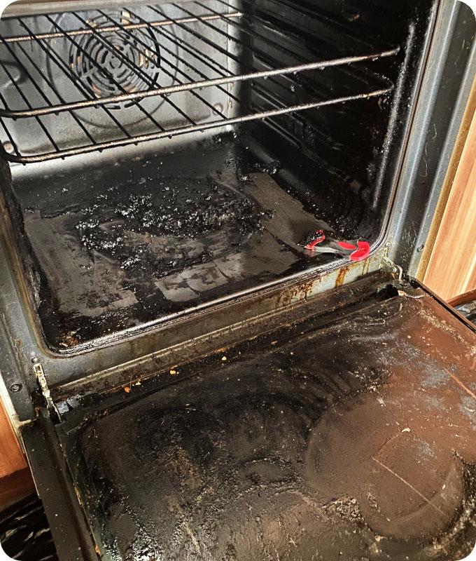 The photo shows the inside of a dirty oven. From the looks of it, this oven hasn’t been cleaned for ages. It is so dirty and gross. The inside surfaces of the oven chamber are caked with layers upon layers of black gunk and burnt grease. The oven door glass is so dirty that it looks completely black, it has lost its transparency entirely. It probably smells quite gross as well.