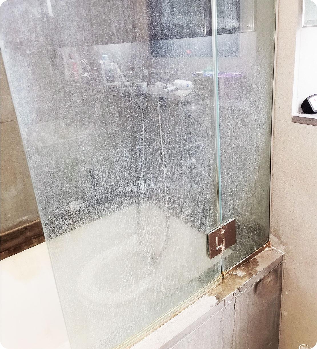 The image shows a bathtub shower cabin that appears quite messy and dirty. The glass door is all smudgy and grainy, covered in soapy scum and limescale.