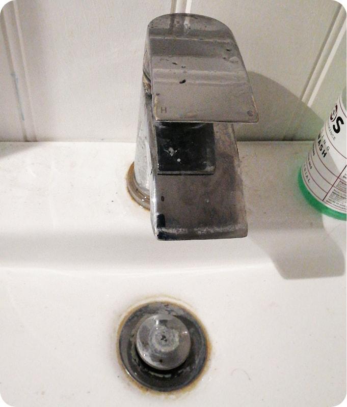 A close-up shot of a dirty white porcelain bathroom sink with a chrome faucet and a chrome drain covered in limescale.