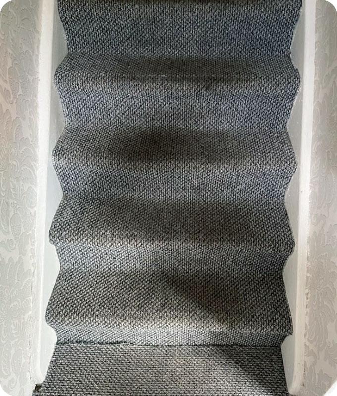Photograph of a dark grey carpeted stairway that appears heavily soiled and in need of deep cleaning. The carpet is covered in visible dirt, dust, and grime, with darker patches of dirt concentrated in high-traffic areas.