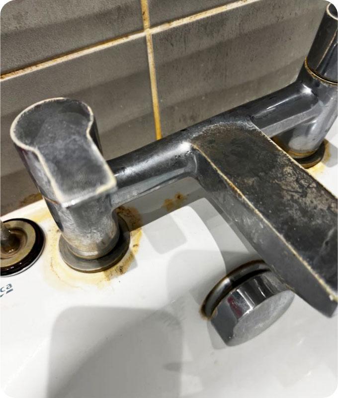 An image of a dirty chrome bathroom faucet covered in grime, hard water stains, and soap scum. The chrome finish appears dull and tarnished, with visible dirt and water spots all over. The faucet handles and spout are coated with soap residue and grime, giving it a neglected and unclean appearance.