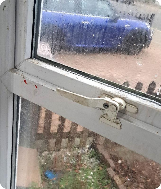 A close shot of a white uPVC window from the inside of a room. The window and the frame are quite dirty, you can barely see through the window. There is dark gunk and grime smudged all over the window frame.