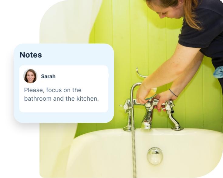 A domestic cleaner wearing a dark blue Fantastic Services uniform is shown in a domestic bathroom. She is standing over a white bathtub and using a sponge to clean the chrome tub faucet. Both the tub and the green tiled wall behind it appear perfectly clean and shiny. There is a segment which has been edited into the photo, showing a note from a customer. The note says “ please focus on the bathroom and the kitchen.”