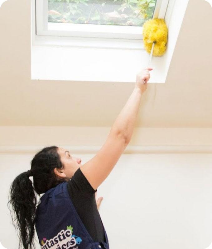 A brightly lit room of a house with skylights. A domestic cleaner who is wearing a dark blue Fantastic Services uniform is reaching up to one of the skylights and wiping off the dust from the white uPVC window frame with a yellow home duster.