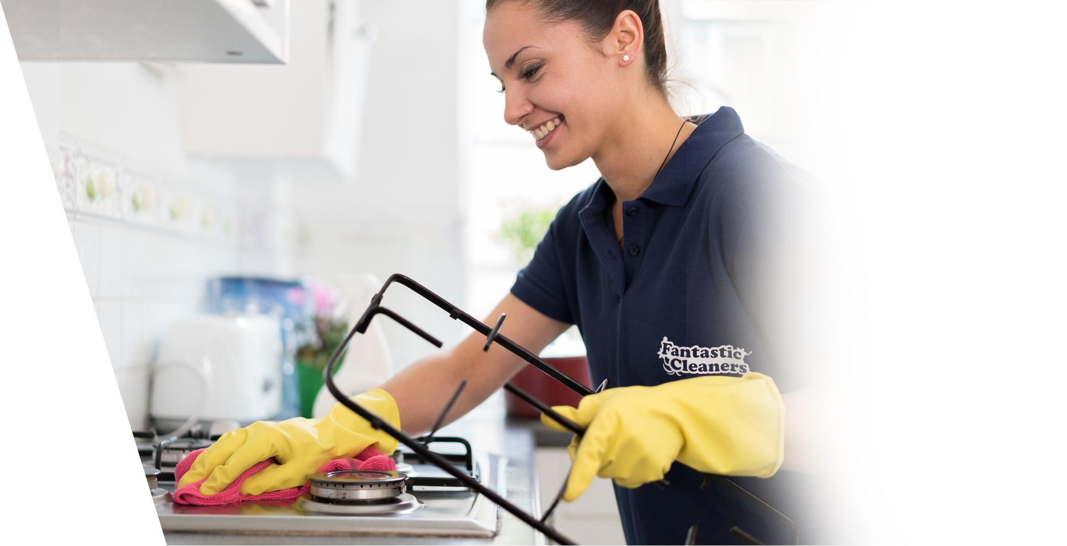 A Fantastic Services professional cleaner who is wearing a dark blue uniform and yellow latex gloves. She is in a domestic kitchen, using a cloth to wipe a stove. The surfaces and the floor of the kitchen appear spotless and shiny.