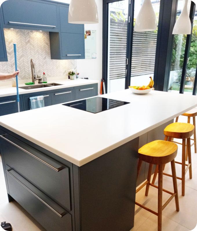 A brightly lit domestic kitchen in the centre of which is a wide counter with an integrated black stove, surrounded by tall wooden chairs without backrests. The kitchen appears very neat and clean, there is a bowl of fruits on the kitchen countertop. The floor of the kitchen and all the various surfaces are shiny and polished clean.