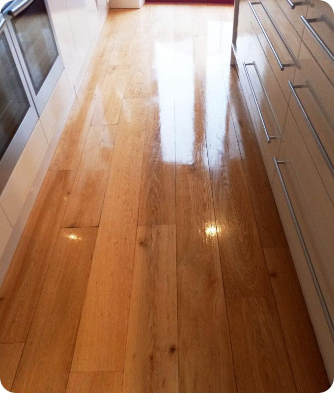A wooden kitchen floor that has been cleaned and polished to perfection. The floor is so shiny and glossy that it is actually reflective of the daylight that comes from the windows.