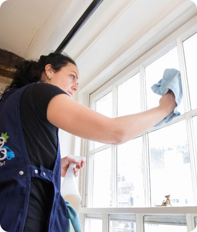 A Fantastic Services domestic cleaner who is wiping a window on the inside in a bright room of a house. She is using a blue wiping cloth to diligently clean the glass.