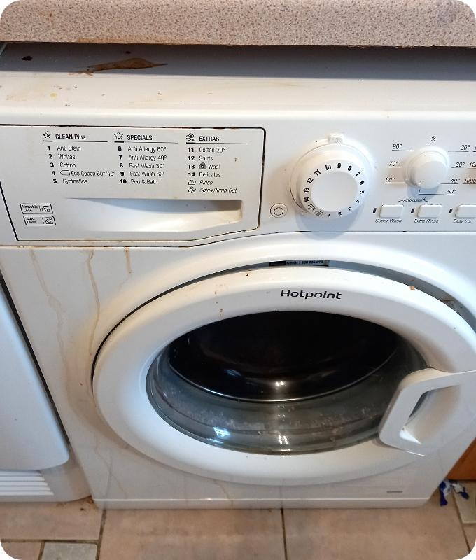 A close shot of a white washing machine positioned under a kitchen counter over a tiled floor. It appears that the appliance's exterior is quite dirty with stains from various spilt substances.