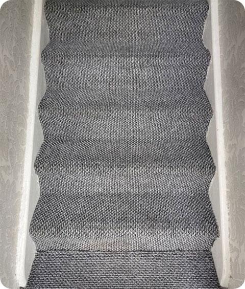 Photograph of a dark grey carpeted stairway that has been professionally cleaned with hot water extraction machines. The carpet appears revitalized, with no visible signs of dirt, dust, or grime.