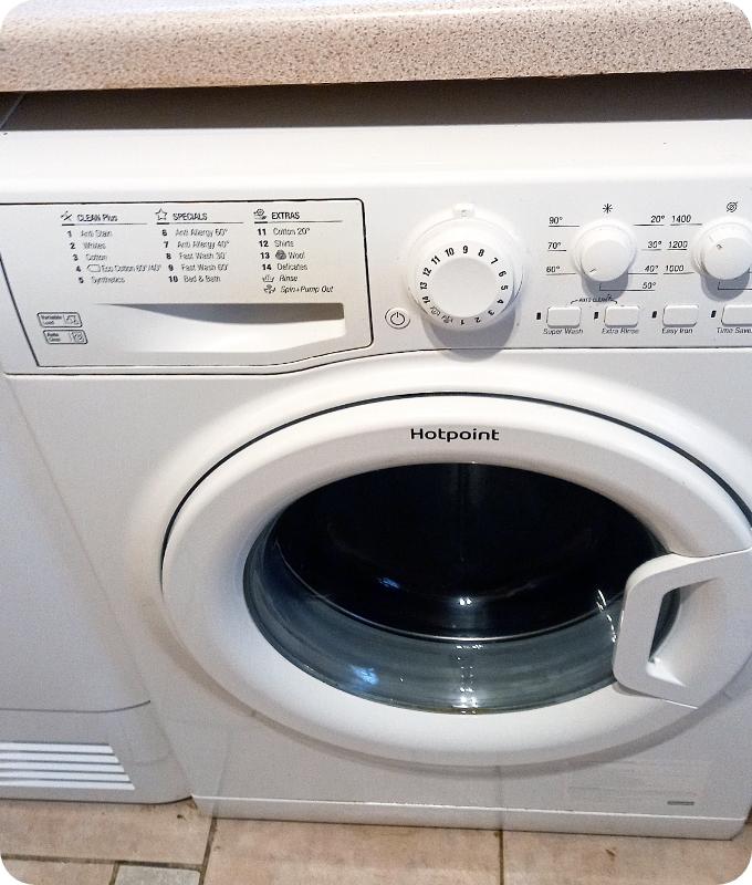 A close shot of the same white washing machine positioned under a kitchen counter over a tiled floor. The washing machine has been wiped clean and the stains from spillage are removed.