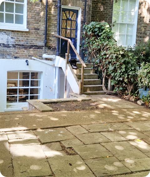 The image shows the same flagstone walkway around a house. Here the weeds between stones have been removed and the shrub has been trimmed.