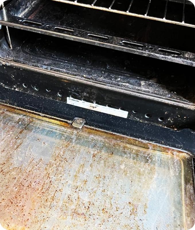 The image shows a close-up of a dirty oven glass. The surface is caked with greasy splatters and crusty dark brown stains.